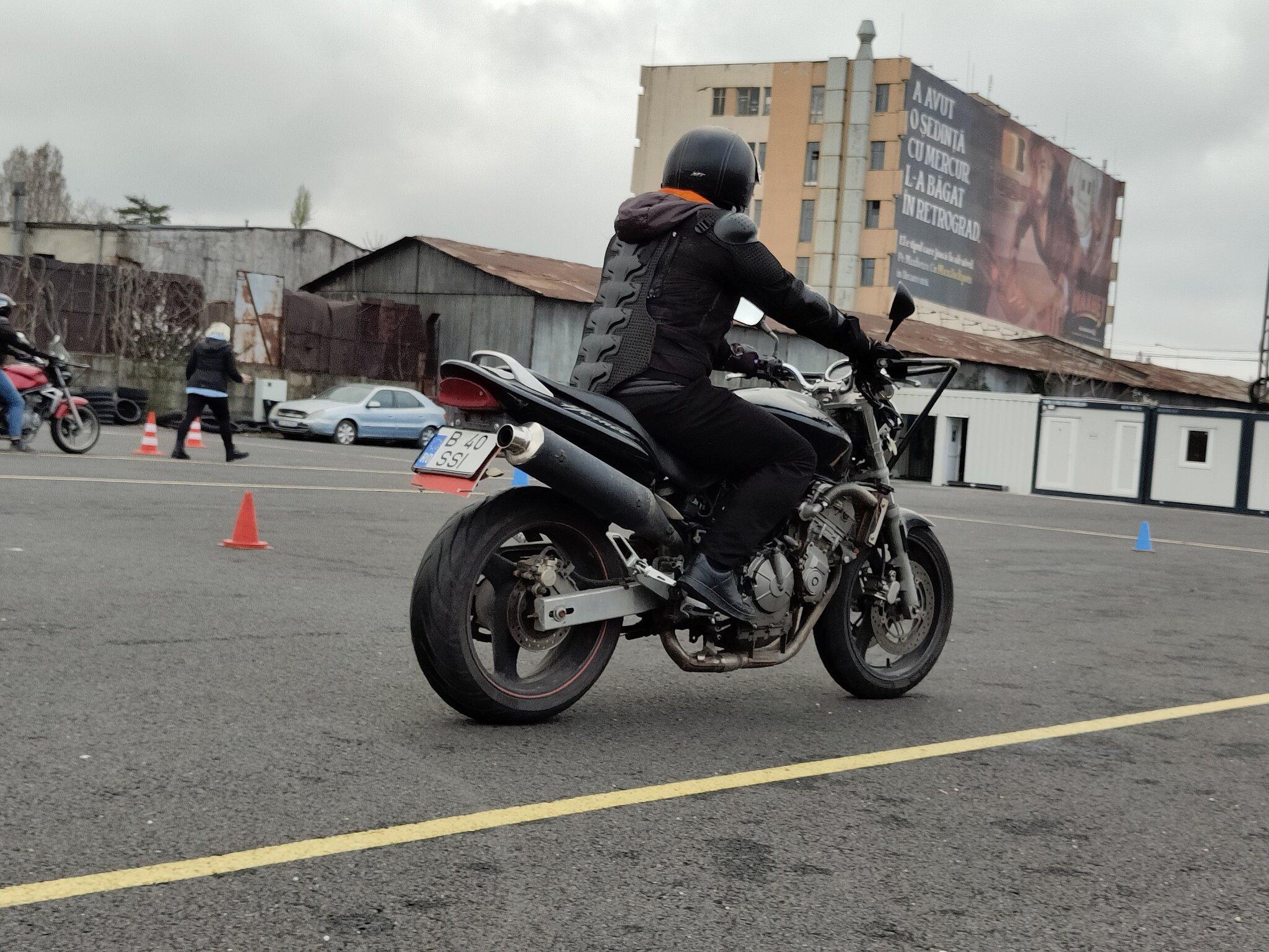 Motociclist exersând opturi pe o motocicletă neagră la poligonul Școlii de Șoferi Giulești, cu instructoarea și un alt cursant în fundal
