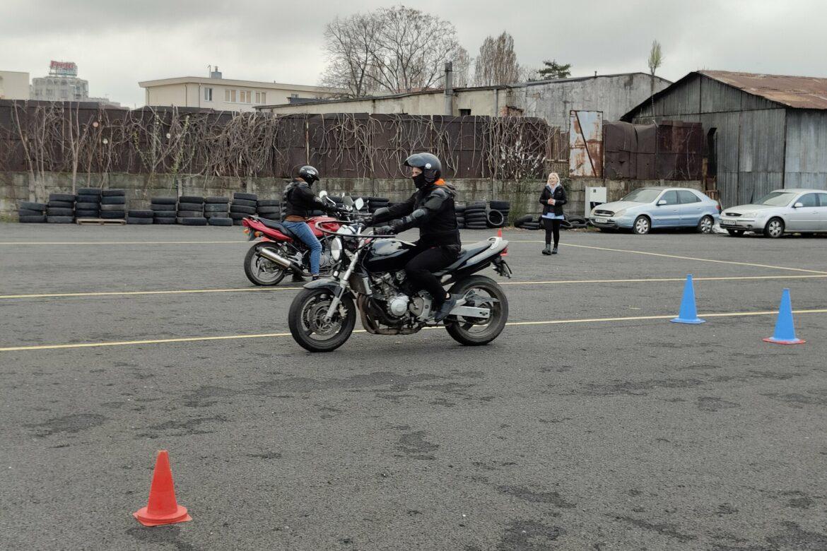 Doi motocicliști exersând pe poligonul Școlii de Șoferi Giulești, cu marcajele vizibile și instructoarea lor supervizând