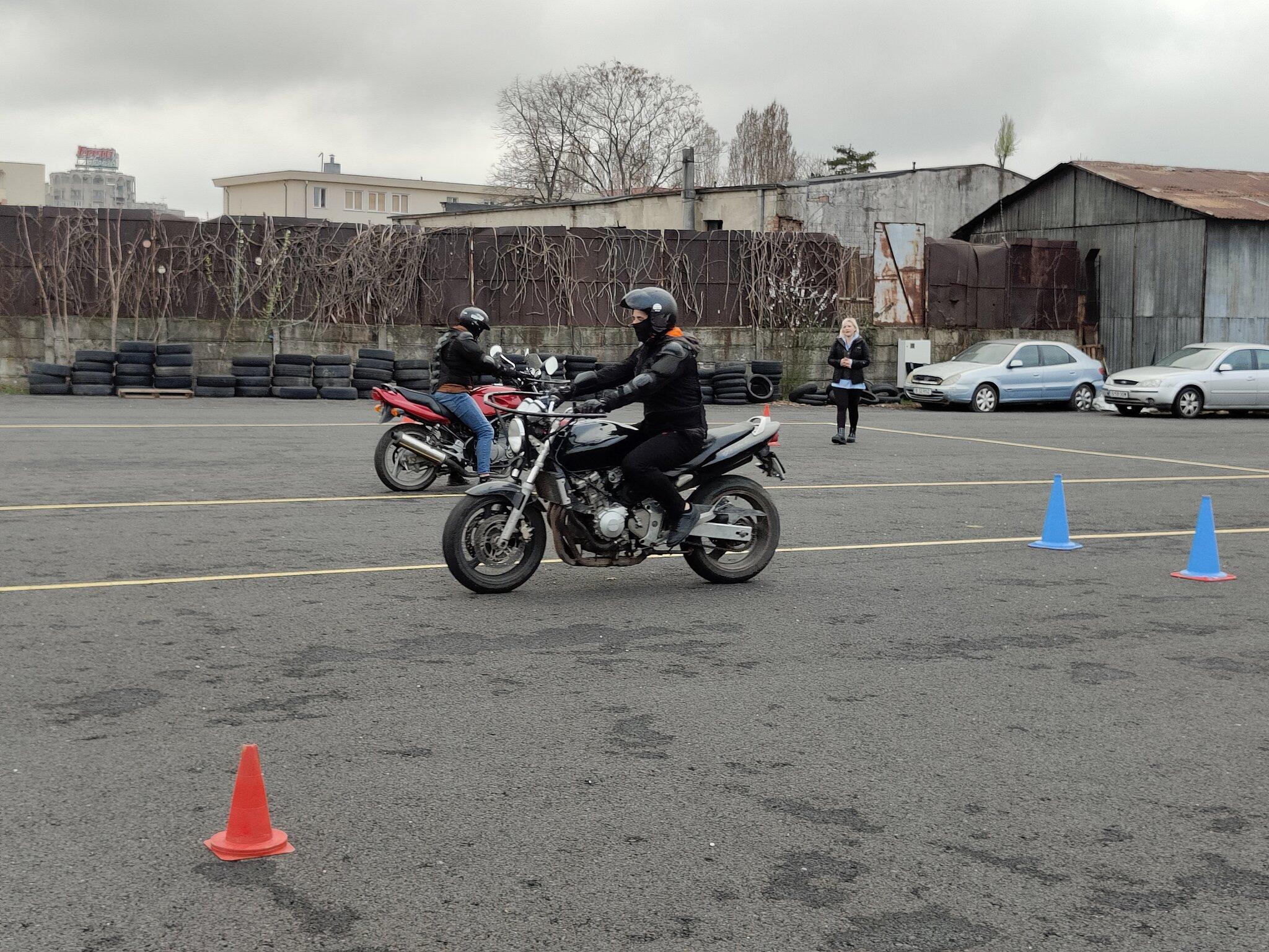 Doi motocicliști exersând pe poligonul Școlii de Șoferi Giulești, cu marcajele vizibile și instructoarea lor supervizând
